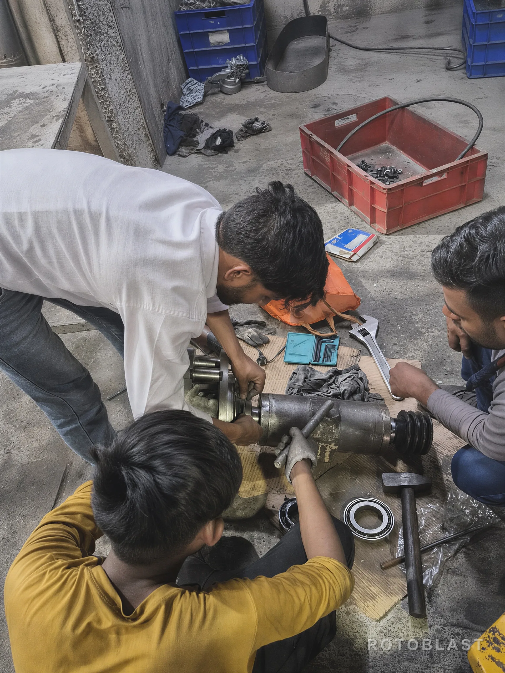 Three maintenance technicians crouched on a workshop floor around a blast wheel assembly stripped down to its shaft, hub and bearing, with spanners, allen keys and a replacement bearing laid out beside them